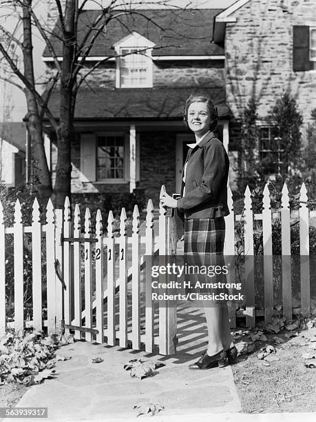 1940s SMILING PRETTY YOUNG TEENAGE GIRL STANDING BY WHITE PICKET FENCE IN FRONT OF STONE HOUSE IN AUTUMN LOOKING AT CAMERA