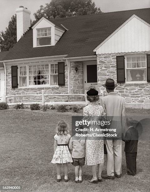 1950s FAMILY OF FIVE WITH BACKS TO CAMERA ON LAWN LOOKING AT FIELDSTONE HOUSE