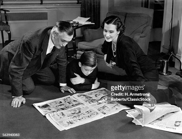 1930s 1940s FATHER AND MOTHER READING SUNDAY NEWSPAPER COMICS WITH SON SITTING ON LIVING ROOM FLOOR TOGETHER