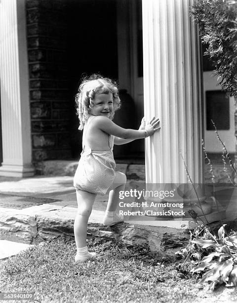 1930s SMILING TODDLER GIRL STEPPING UP ONTO PORCH TOUCHING COLUMN SUMMER BLOND CURLY HAIR HOUSE LAWN