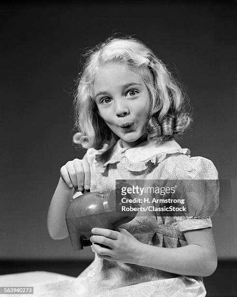 1940s CHILD BLOND GIRL PUTTING MONEY COIN INTO PIGGY BANK LOOKING AT CAMERA