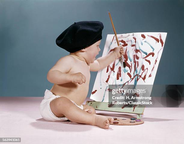 1960s BABY ARTIST WEARING BLACK BERET SITTING IN FRONT OF EASEL PAINTING