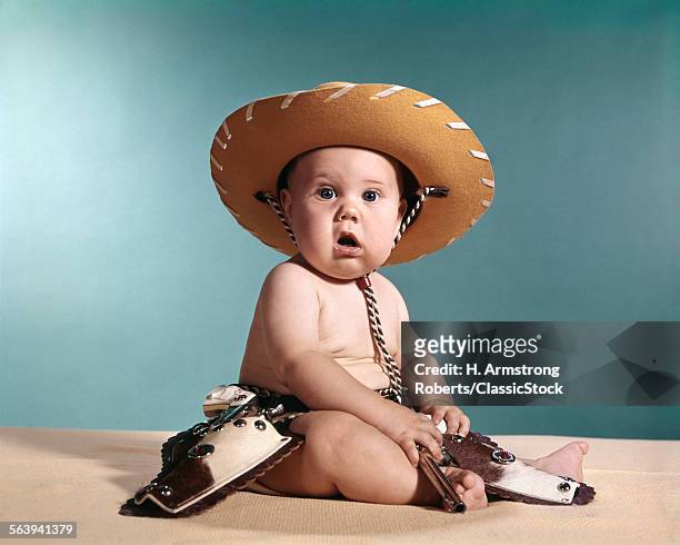 1960s BABY WEARING COWBOY COSTUME WITH FUNNY FACIAL EXPRESSION LOOKING AT CAMERA