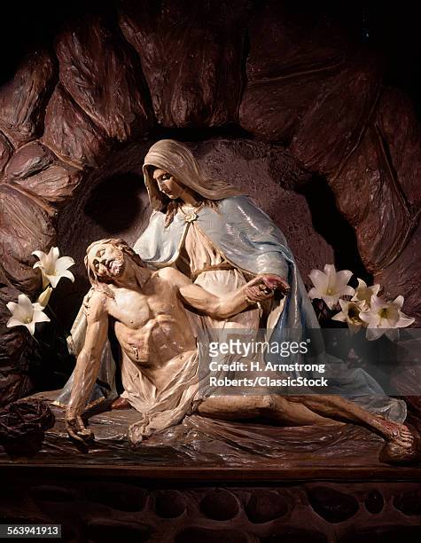 A PIETA STATUE OF MARY MOTHER HOLDING DEAD BODY OF JESUS CHRIST IN HER LAP WITH WHITE LILIES