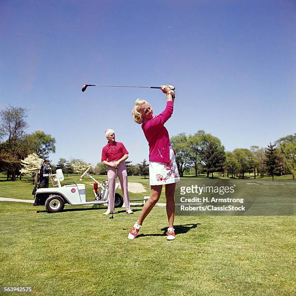 1960s 1970s MATURE WOMAN SWINGING GOLF CLUB AS MAN STANDS BY GOLF CART
