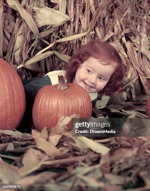 1950s CUTE LITTLE GIRL WITH RED HAIR HIDING BEHIND PUMPKIN SMILING LAUGHING DRIED CORN STALK HARVEST AUTUMN HALLOWEEN