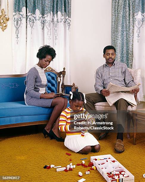 1960s AFRICAN AMERICAN FAMILY MOTHER FATHER SITTING READING LIVING ROOM GIRL DAUGHTER PLAYING ON FLOOR WITH LEGO TOYS