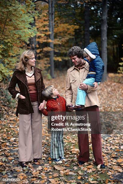 1970s FAMILY OF FOUR WALKING IN AUTUMN LANDSCAPE HOLDING HANDS FATHER CARRYING GIRL TALKING TO BOY