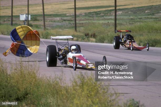 1970s 1980s DRAG RACE TWO ALCOHOL BURNING FUEL DRAGSTERS ON STRIP NEAR FINISH OPEN DROGUE PARACHUTE DEPLOYED SLOWING WINNER