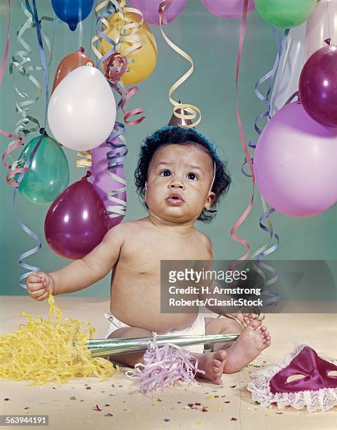 1960s AFRICAN AMERICAN BABY BOY WEARING PARTY HAT AMID BALLOONS AND STREAMERS NEW YEARS NOISEMAKER AND MASK