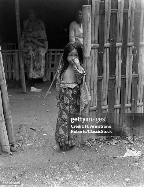 1920s 1930s LITTLE JAVANESE GIRL IN BATIK SKIRT STANDING BY BAMBOO FENCE HAND UP TO FACE JAVA INDONESIA
