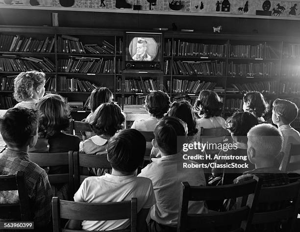 1950s VIEW FROM BEHIND GROUP OF ANONYMOUS CHILDREN IN ELEMENTARY SCHOOL LIBRARY WATCHING TELEVISION TV SCREEN