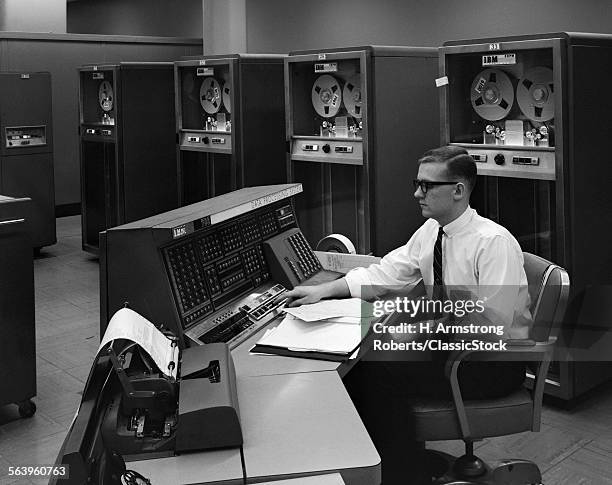 1960s SIDE VIEW OF MAN IN SHIRT TIE & THICK BLACK GLASSES WORKING WITH IBM DATA PROCESSING SYSTEM