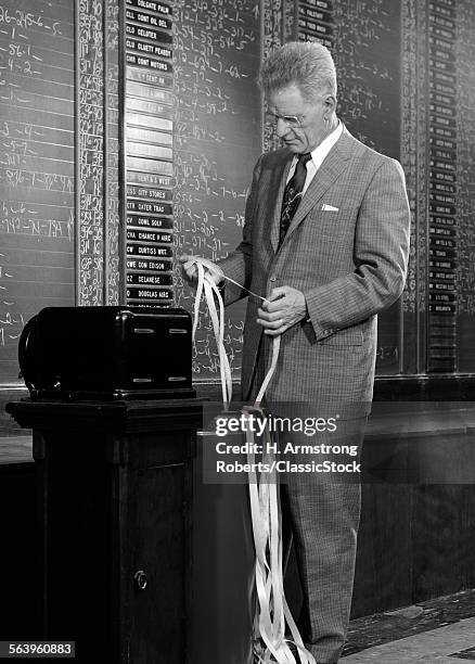 1950s 1960s SERIOUS BUSINESSMAN STOCK BROKER READING QUOTATION TAPE AT OFFICE TICKER-TAPE MACHINE ON EXCHANGE INDOOR