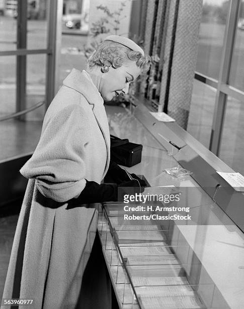 1950s STYLISH WOMAN WEARING HAT OVERCOAT AND GLOVES WRITING DEPOSIT OR WITHDRAWAL SLIP IN BANK BRANCH