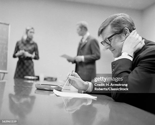 1970s WORRIED BUSINESSMAN SEATED CONFERENCE TABLE TWO PEOPLE IN BACKGROUND