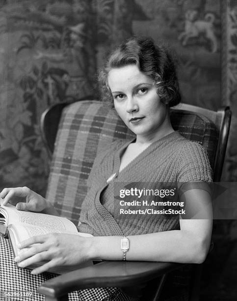 1930s WOMAN SITTING IN PLAID ARMCHAIR HOLDING BOOK LOOKING AT CAMERA