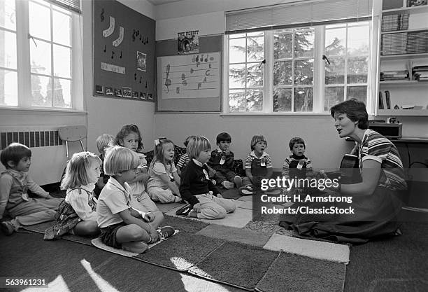 1980s GRADE SCHOOL TEACHER SITTING ON FLOOR WITH STUDENTS PLAYING GUITAR & SINGING