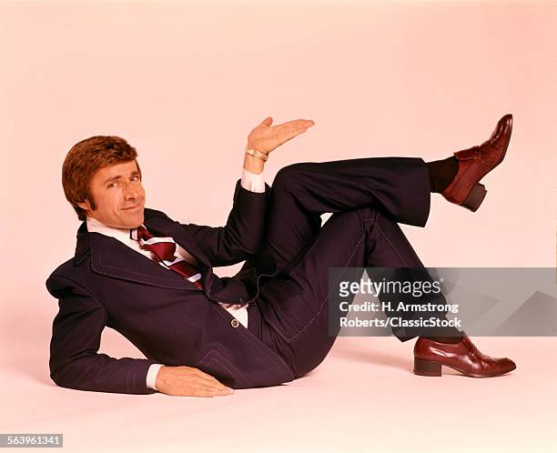 1970s SMILING MAN IN BUSINESS SUIT AND TIE LYING DOWN LEGS CROSSED LOOKING AT CAMERA MAKING UPLIFTING GESTURE WITH HAND