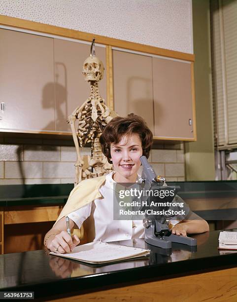 1960s GIRL PRE-MED STUDENT WHITE BLOUSE NOTEBOOK SEATED AT MICROSCOPE IN CLASSROOM LAB LOOKING AT CAMERA SKELETON IN BACKGROUND