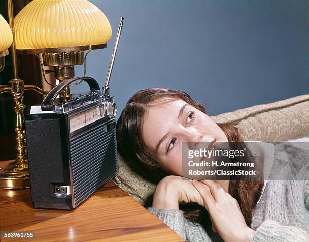 1960s TEENAGE GIRL LYING ON COUCH LISTENING TO MUSIC ON PORTABLE BATTERY POWERED RADIO