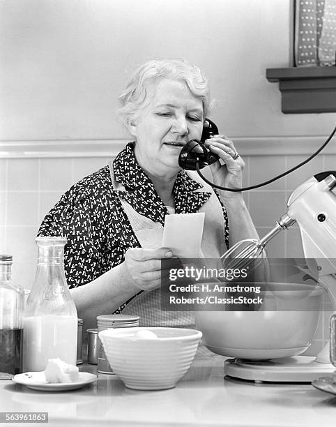 1940s WOMAN GRANDMOTHER HOUSEWIFE COOK IN KITCHEN WEARING APRON WITH FOOD MIXER READING INGREDIENTS LIST TALKING ON TELEPHONE