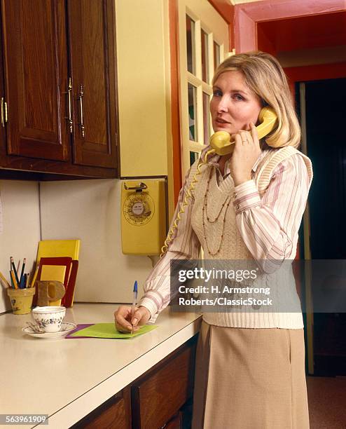 1970s SERIOUS BLOND WOMAN HOUSEWIFE TALKING ON YELLOW WALL PHONE WRITING ON NOTE PAD ON KITCHEN COUNTER LOOKING AT CAMERA