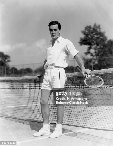 1930s MAN WEARING TENNIS WHITES STANDING LOOKING AT CAMERA HOLDING RACKET HANDS ON THE NET