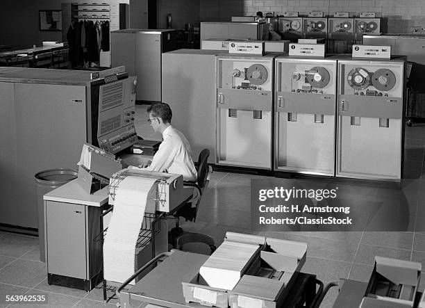 1960s MAN PROGRAMMING LARGE MAINFRAME COMPUTER SURROUNDED BY DATA TAPE DRIVES INDOOR