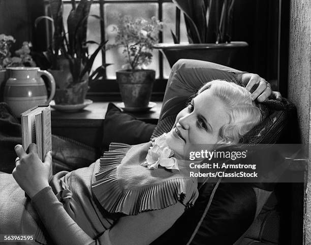 1930s BLOND WOMAN LYING BACK IN WINDOW SEAT WITH PILLOWS HOLDING BOOK IN ONE HAND