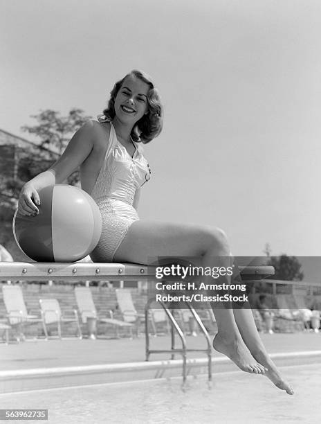 1950s BATHING BEAUTY IN SATIN ONE-PIECE SWIMSUIT LOOKING AT CAMERA SITTING DIVING BOARD HOLDING BEACH BALL