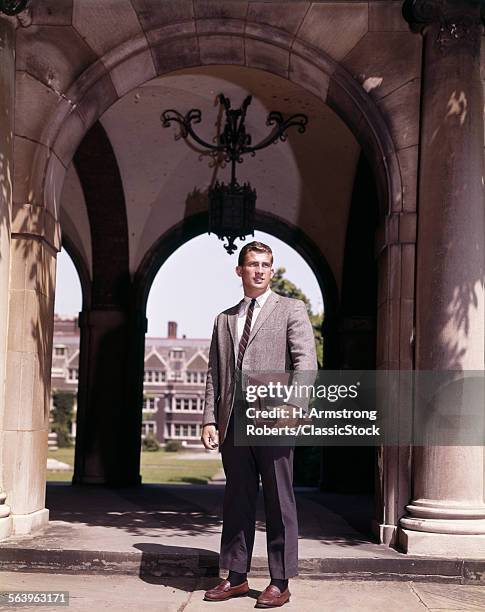 1960s MAN COLLEGE STUDENT STANDING NEAR ARCH LEADING TO COLLEGE CAMPUS QUADRANGLE
