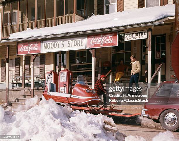 1960s TWO MEN PUMPING GAS FOR SNOWMOBILE ON TRAILER PARKED BY FRONT PORCH OF GENERAL STORE AND POST OFFICE EAST DOVER VT USA