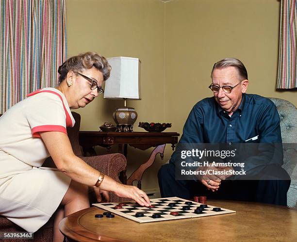 1970s SENIOR COUPLE MAN AND WOMAN SITTING IN LIVING ROOM PLAYING CHECKERS