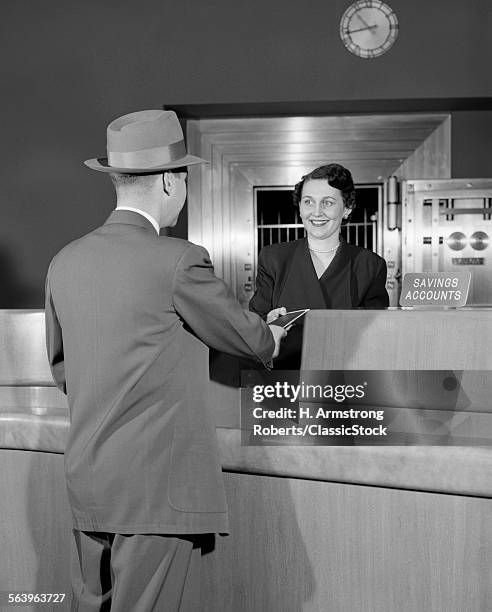 1950s MAN IN SUIT AND HAT HANDING TRANSACTION TO SMILING WOMAN TELLER OPEN BANK VAULT IN BACKGROUND