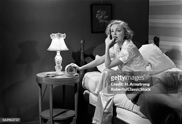 1950s WOMAN SILK PAJAMAS SITTING EDGE OF BED STRESSED SERIOUS WORRIED SLEEPLESS HOLDING ALARM CLOCK NIGHT TABLE LAMP INSOMNIA