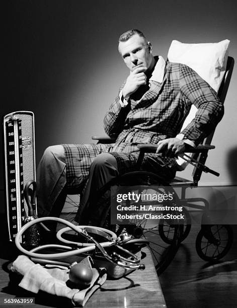 1950s PENSIVE WORRIED MAN IN ROBE SEATED IN WHEELCHAIR IN MOODY SETTING WITH BLOOD PRESSURE METER AND STETHOSCOPE IN FOREGROUND