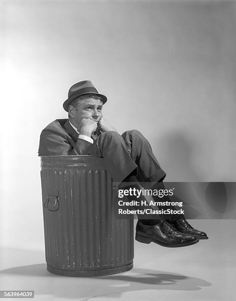 1960s BUSINESSMAN IN SUIT AND HAT SITTING IN TRASHCAN