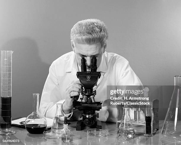 1950s MAN IN LAB COAT SITTING AT TABLE LOOKING THROUGH MICROSCOPE SURROUNDED BY VARIOUS BEAKERS TEST TUBES AND PETRI DISHES