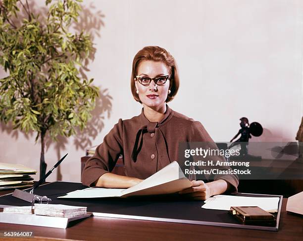 1960s WOMAN BUSINESS EXECUTIVE WITH SERIOUS EXPRESSION WEARING GLASSES SEATED BEHIND DESK LOOKING AT CAMERA