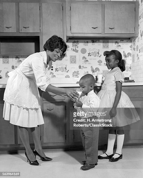 1960s SMILING AFRICAN AMERICAN WOMAN MOTHER IN APRON AND PUMPS GIVING A GLASS OF MILK TO SON AND DAUGHTER WITH MARY JANE SHOES