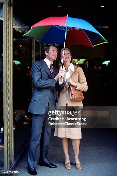 1970s PROFESSIONAL MAN WOMAN COUPLE STANDING TOGETHER UNDER RAIN UMBRELLA