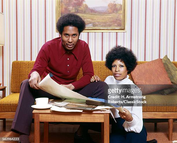 1970s AFRICAN AMERICAN MAN WOMAN COUPLE HUSBAND WIFE LOOKING AT CAMERA EXAMINING NEW HOUSE BLUEPRINTS