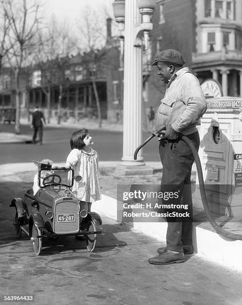 1920s SMILING LITTLE GIRL STANDING BESIDE HER PEDAL CAR ASKING FOR GASOLINE FROM AFRICAN AMERICAN GAS PUMP ATTENDANT