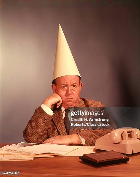 1960s SAD DEPRESSED BUSINESSMAN DESK WEARING DUNCE CAP MAN MEN BUSINESS TEACHER MISTAKE ERROR WRONG FUNNY SILLY