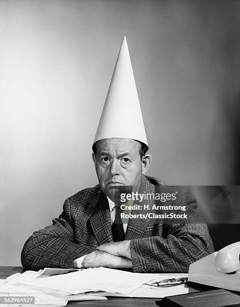 1960s UNHAPPY BUSINESSMAN BEHIND DESK WEARING DUNCE CAP LOOKING AT CAMERA