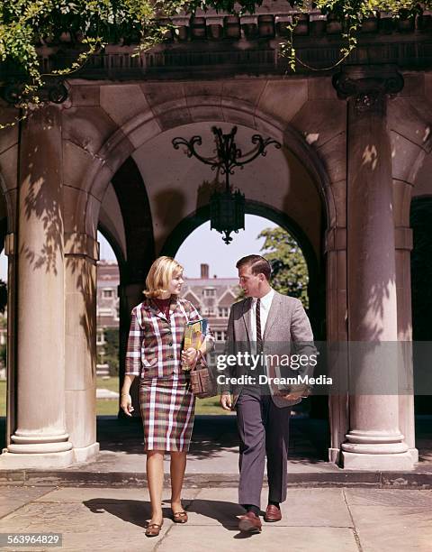 1960s MAN AND WOMAN STUDENTS WITH BOOKS AT ARCH LEADING TO CAMPUS QUADRANGLE