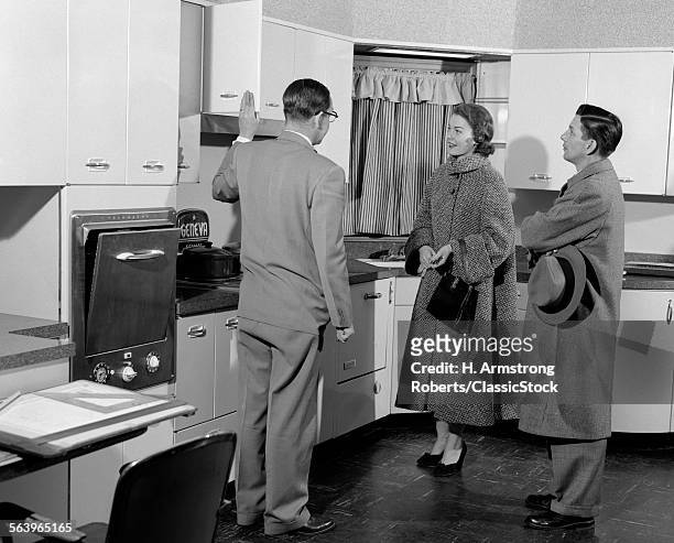 1950s SALESMAN IN MODEL KITCHEN WITH COUPLE