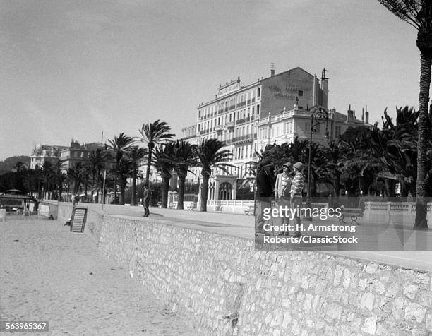 1920s TWO FASHIONABLY DRESSED WOMEN AND A MAN STANDING TALKING ON PROMENADE CANNES FRENCH RIVIERA BY MEDITERRANEAN OCEAN BEACH