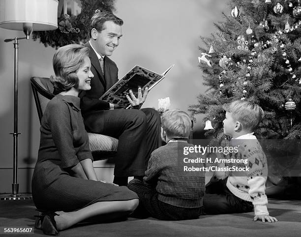 1960s FAMILY FATHER MOTHER TWO SONS SMILING SITTING BY CHRISTMAS TREE IN LIVING ROOM READING A BOOK